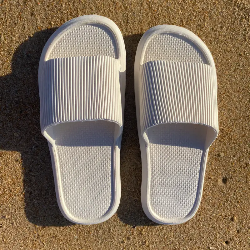 A pair of Reusable Sliders in near-white recycled EVA, set down side by side on sunlit sand, casting a single hard shadow.